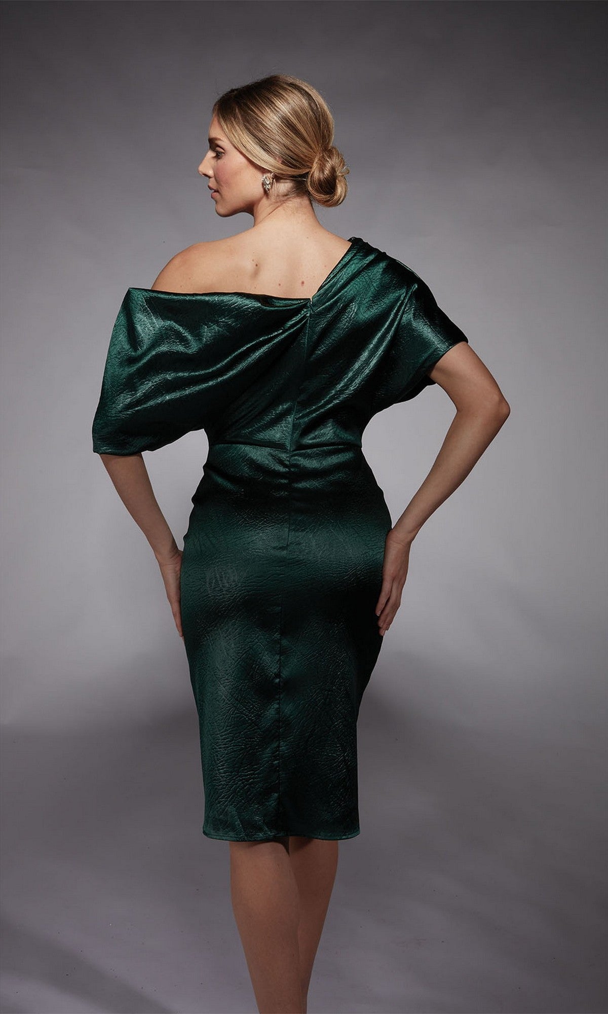 A woman stands with her back to the camera, one hand on her hip, wearing the Alyce 70060 knee-length semi-formal dress in shiny dark green charmeuse against a neutral gray background.
